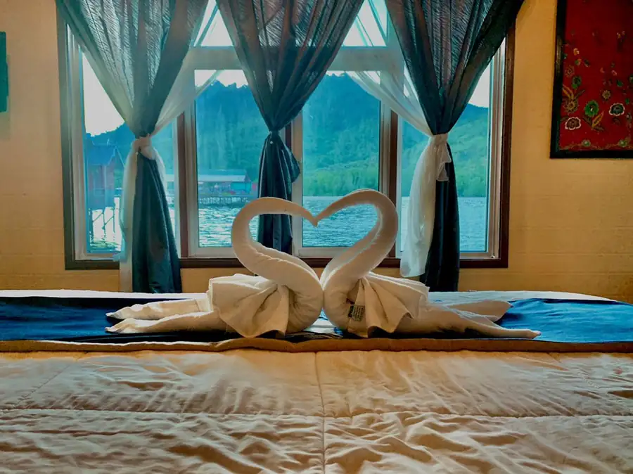 A close-up of a bed adorned with towel swans, positioned in front of a window that frames a beautiful view of calm waters and green hills, capturing authentic local charm at Parapat Water Village Resort.