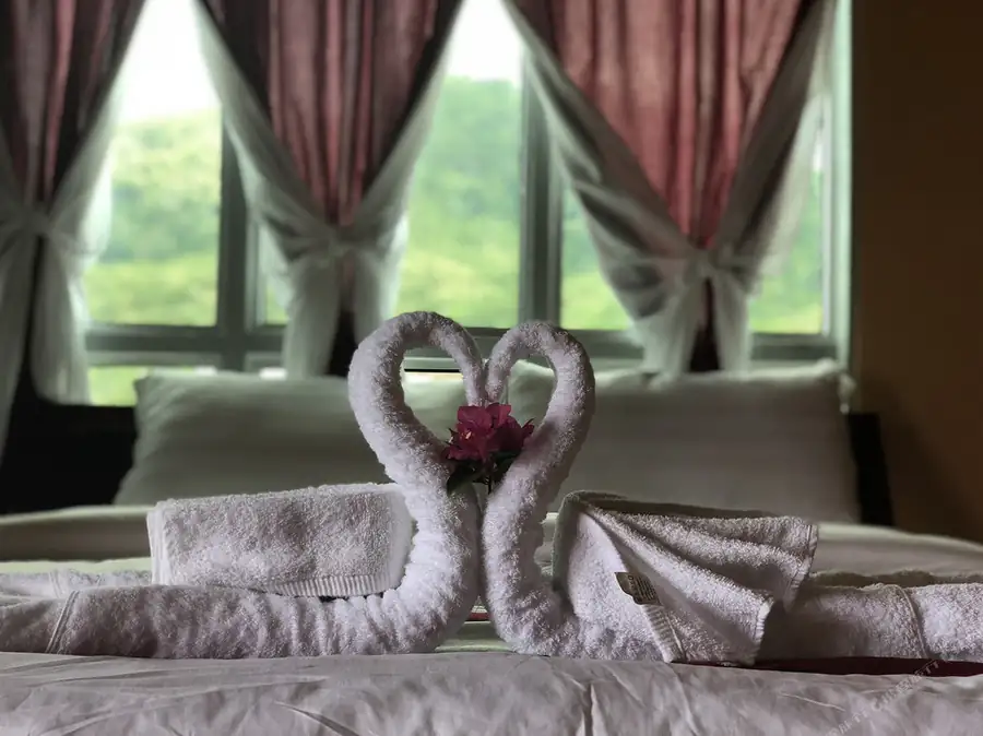 A beautifully arranged bed displaying towel swans embracing a pink flower, set against a bright window with tied-back curtains, illustrating welcoming island hospitality at Parapat Water Village Resort.